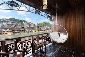 A wooden balcony with a hanging chair, a table, and a view of a riverside town. - Floral Hotel ·Fenghuang Grand Riverview (Fenghuang)