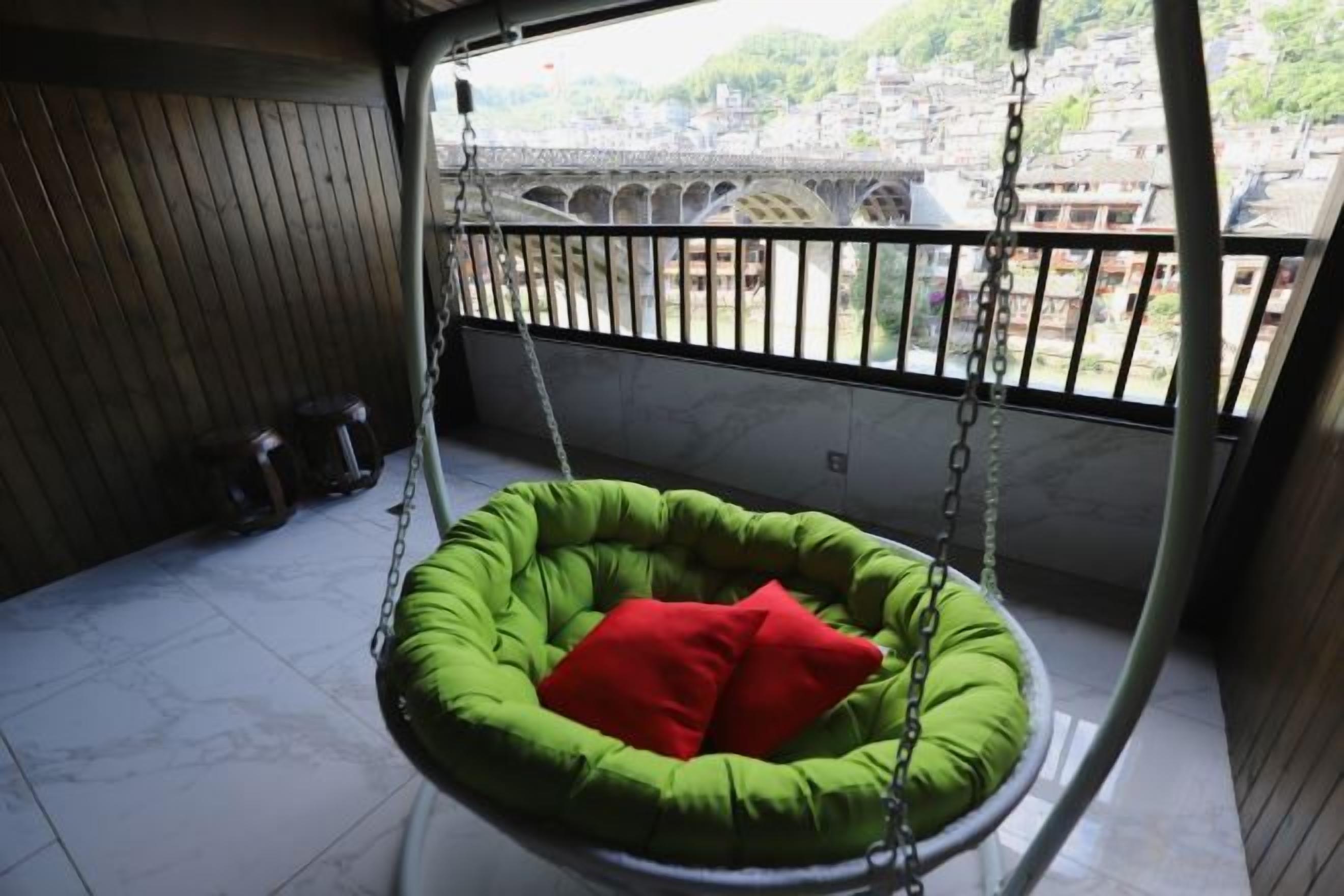 A green hanging chair with a red cushion on a balcony with a view of a bridge and buildings.