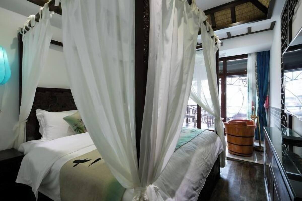 A bedroom with a bed, white curtains, a wooden headboard, and a view of the outside.