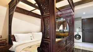Ein Bett mit Baldachin und aufwendigen Schnitzereien, eine Waschmaschine und ein dunkler Holzschrank in einem Raum mit Fliesenboden.