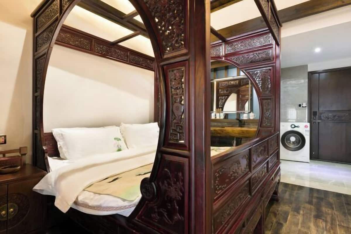 A four-poster bed with intricate carvings, a washing machine, and a dark wooden wardrobe in a room with a tiled floor.