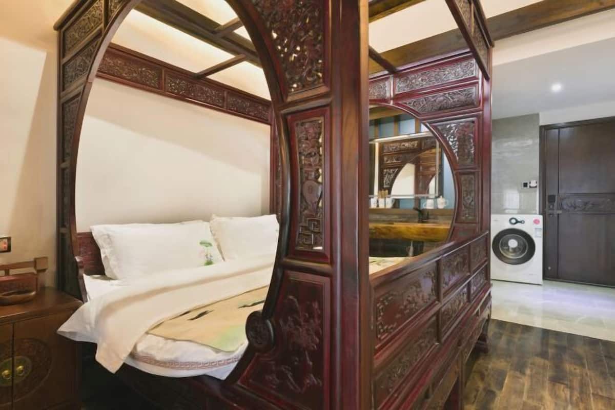 A four-poster bed with intricate carvings, a washing machine, and a dark wooden door in a room with a tiled floor.