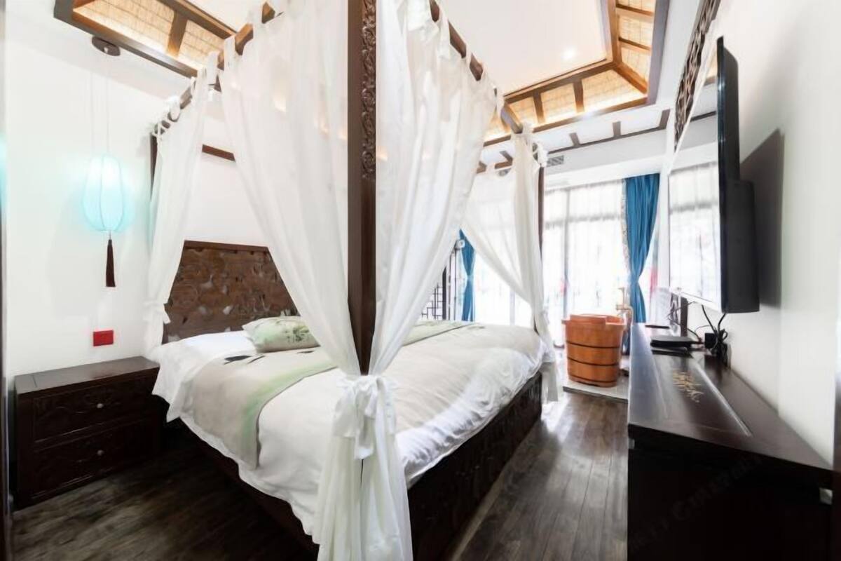 A bedroom with a canopy bed, a wooden headboard, a bedside table, a TV, and a window with curtains.