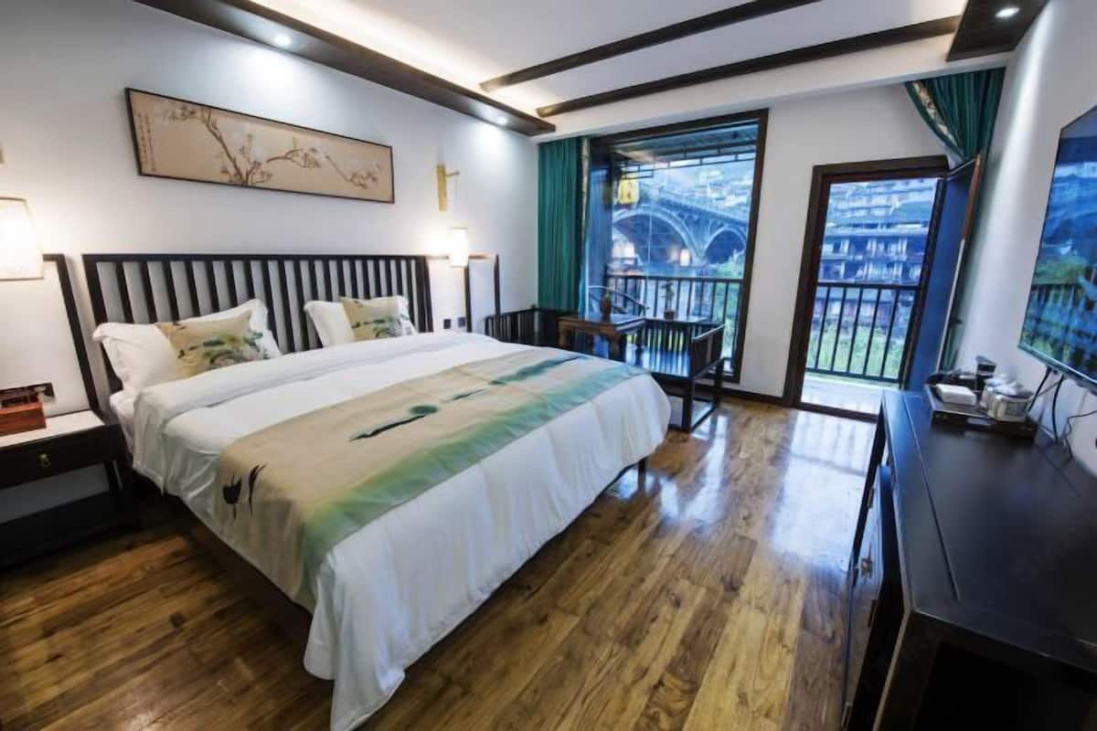 A hotel room with a bed, bedside tables, a desk, and a balcony with a view of a bridge.
