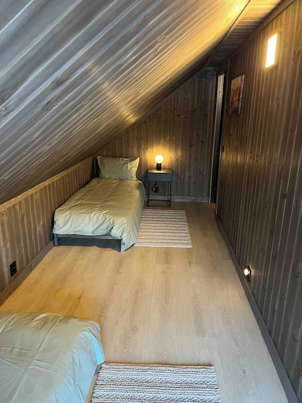 A compact, sloped-roof bedroom with two beds, a bedside table with a lamp, and a wooden floor.