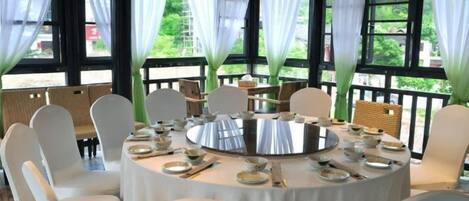 Mesa redonda dispuesta para comer, rodeada de sillas, ventanas amplias con cortinas y vistas al exterior con vegetación.