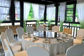 Een ronde eettafel met stoelen eromheen, grote ramen met gordijnen en uitzicht op groen buiten.