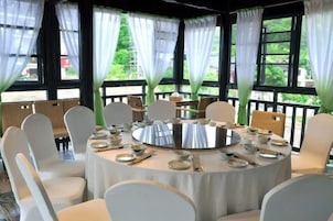 Mesa redonda dispuesta para comer, rodeada de sillas, ventanas amplias con cortinas y vistas al exterior con vegetación.