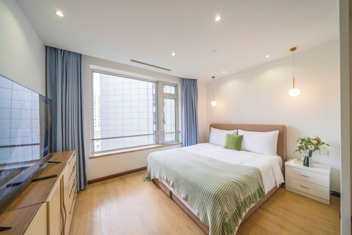 Et moderne hotelværelse med et stort vindue, en seng med en grøn pude, et natbord med en vase blomster og et fjernsyn på en trækommode.