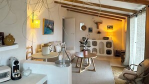 A cozy living room with a sofa, coffee table, and a dining area with chairs. There is a fireplace with decorative items and a large window with curtains. - La Cabane de Gordes (Gordes)