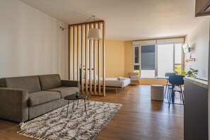 A living room with a sofa, coffee table, and a patterned rug. There is a bed with a wooden partition, a dining area with chairs, and a window with blinds.