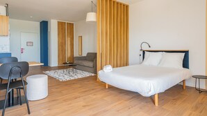 A modern hotel room with a large bed, a seating area with a chair and ottoman, a small table, and a wooden partition. - Ecla Genève Archamps (Archamps)
