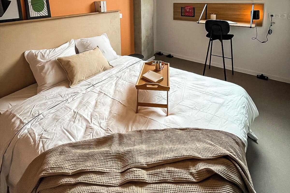 Een slaapkamer met een groot bed, een houten nachtkastje, een zwarte stoel en een raam met uitzicht over de stad.