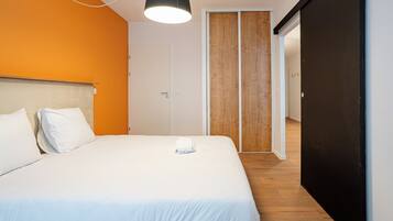 A hotel room with a bed, a white pillow, a white blanket, a black ceiling light, a wooden door, and a wooden floor.