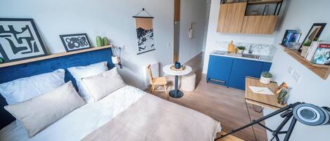 Habitación de hotel compacta con cama, una mesita, una silla, kitchenette y una lámpara de techo.
