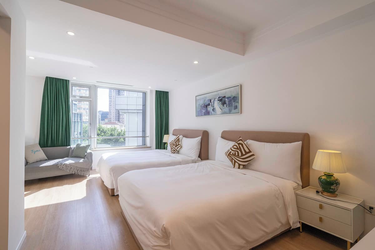 A hotel room with two beds, a sofa, a window with curtains, a painting on the wall, and a bedside lamp.