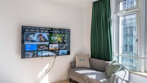 A modern living room with a flat-screen TV mounted on the wall displaying various movie posters. A grey sofa with a cushion labeled "Tym's" is positioned near a window with green curtains. - Tyms Xuhui Spacious 1BR APT  (Shanghai)
