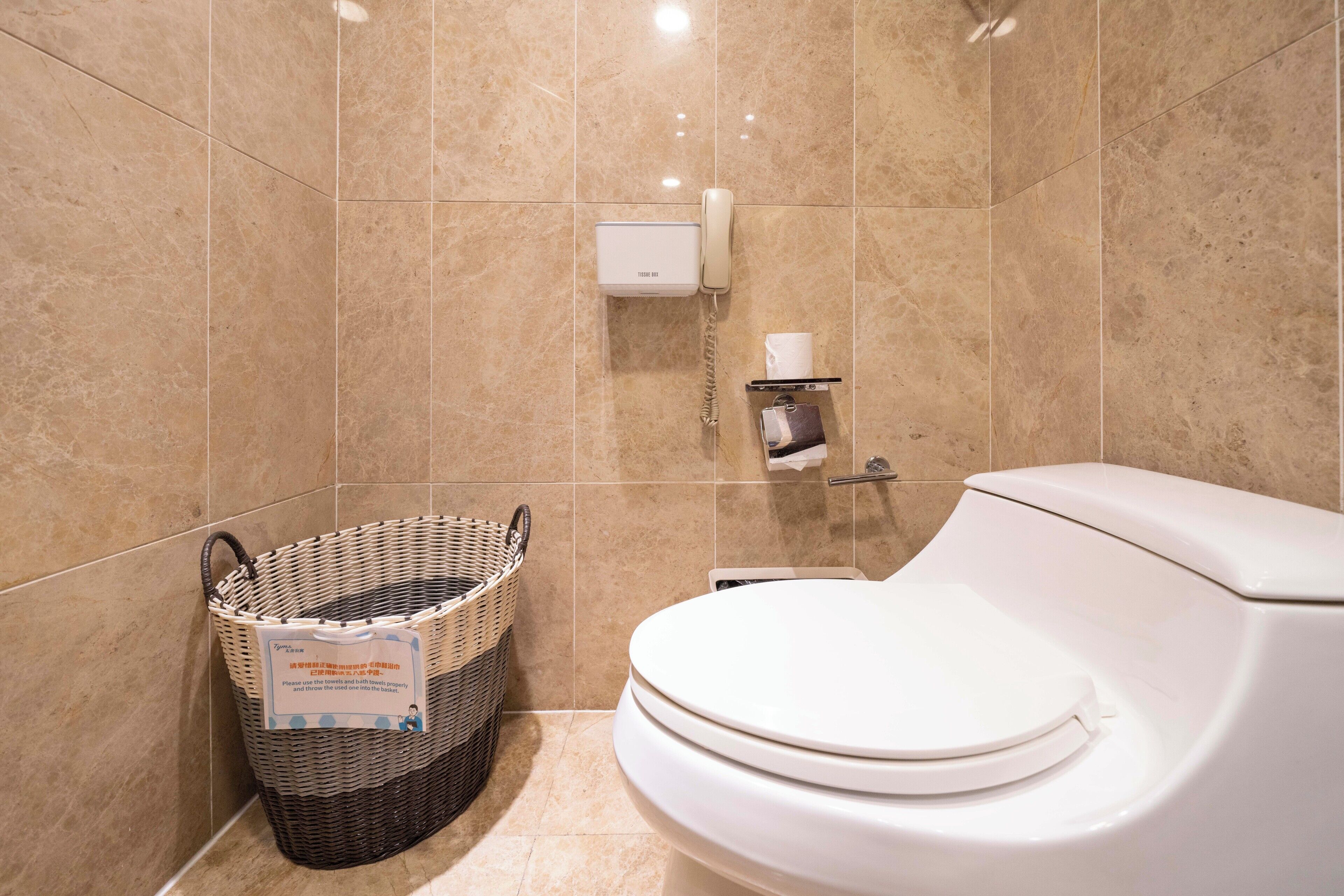 Un baño con inodoro blanco, cesta de mimbre para ropa, dispensador de toallas de papel y teléfono.