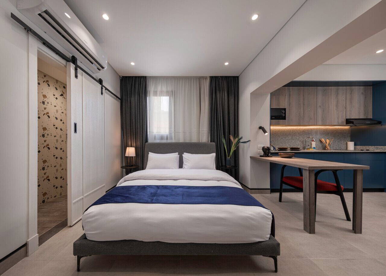 A modern hotel room with a large bed, a small desk, and a kitchenette.