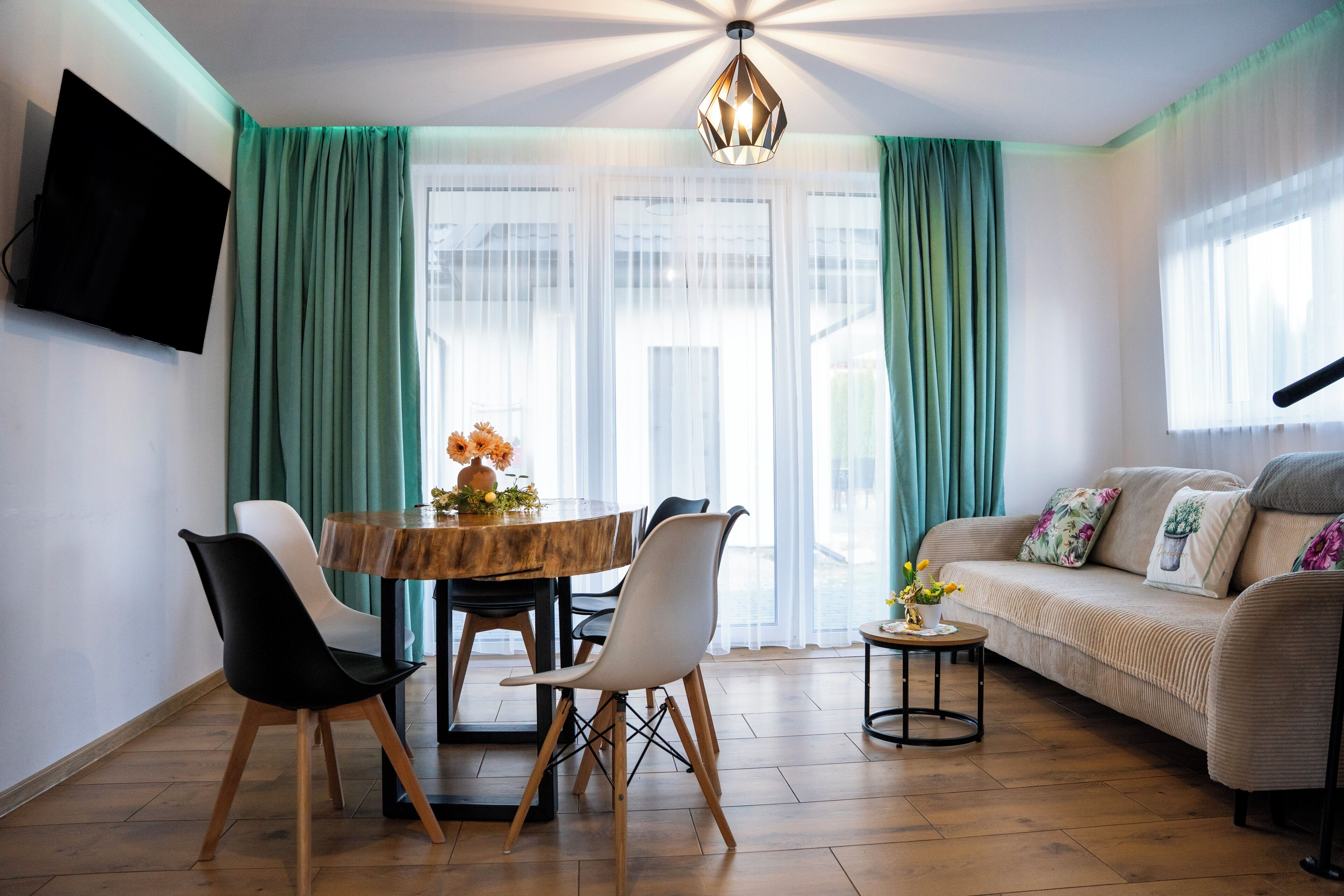 Ein modernes Wohnzimmer mit einer Couch, einem Esstisch mit Stühlen, einem an der Wand montierten Fernseher und einer Deckenleuchte.