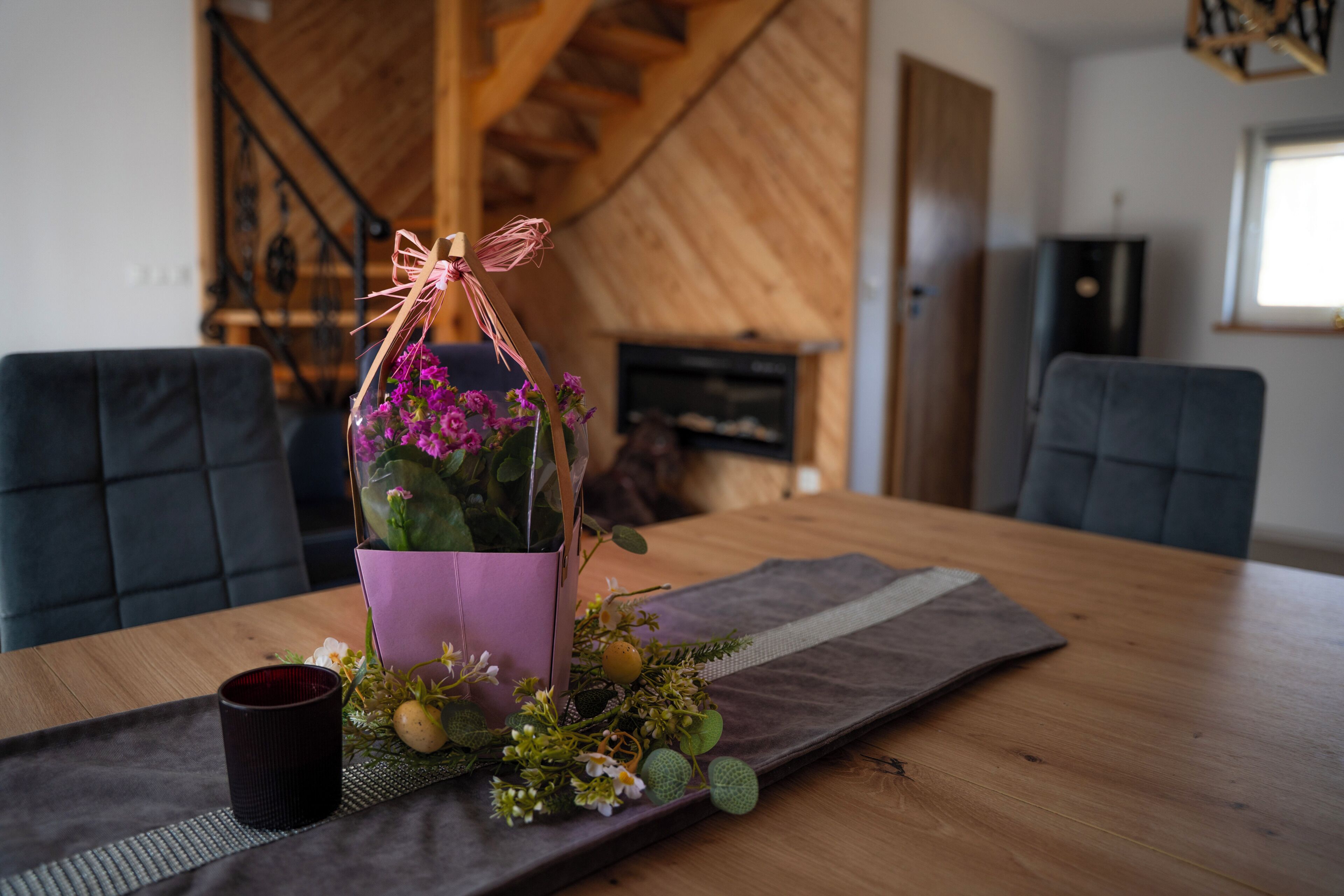 Une table à manger ornée d’un vase violet contenant des fleurs, d’une tasse foncée et d’un set de table en fibres tressées.