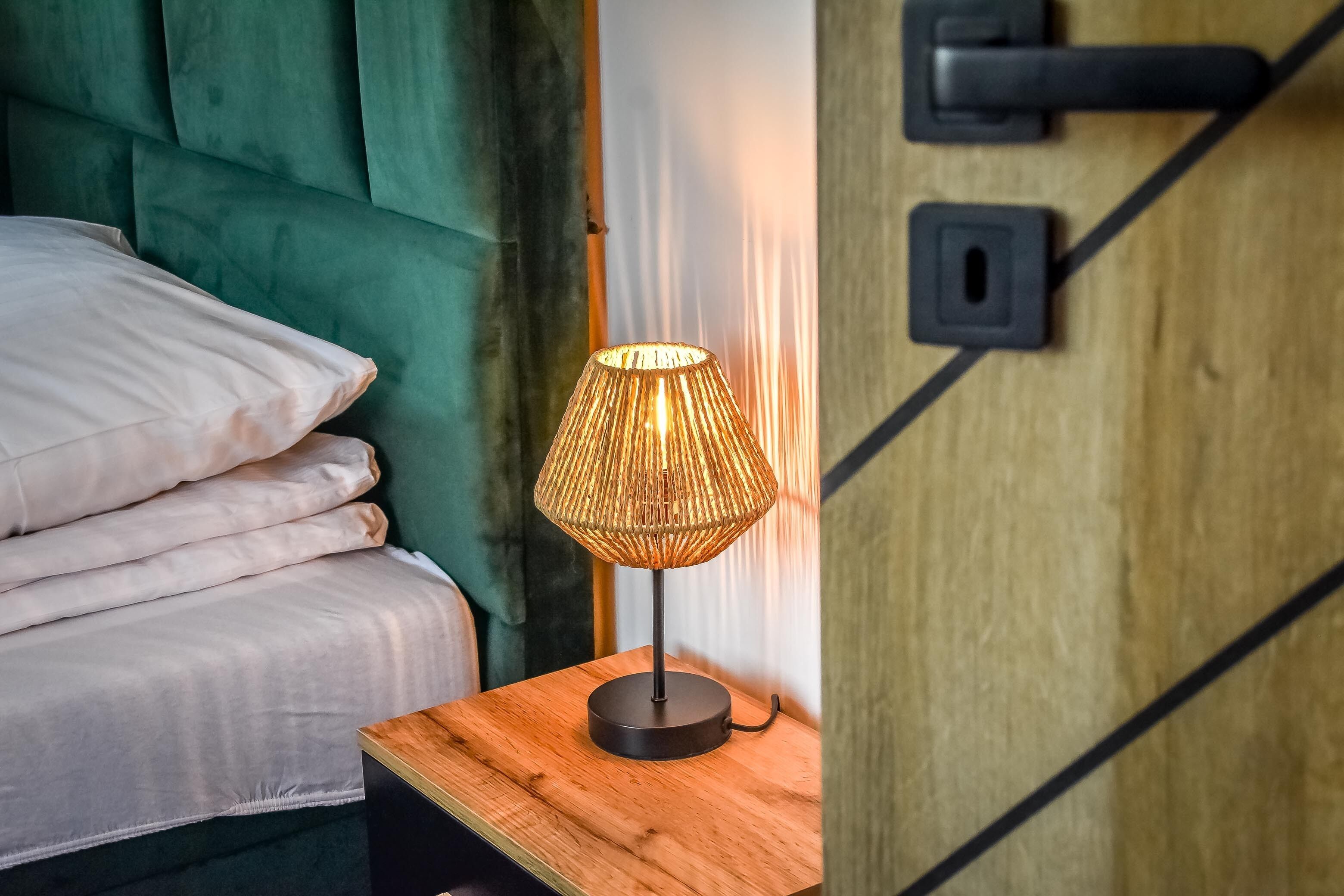 Un lit bien fait, avec une tête de lit verte, une table de chevet en bois surmontée d’une lampe en osier et une porte en bois dotée d’une poignée noire.