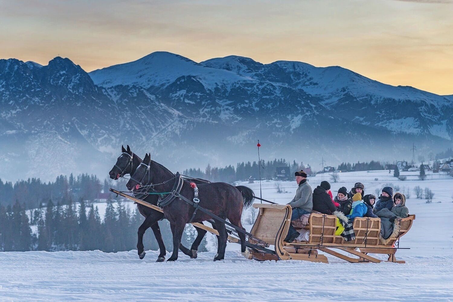 Um trenó puxado a cavalo transporta passageiros por uma paisagem nevada, com montanhas ao fundo.