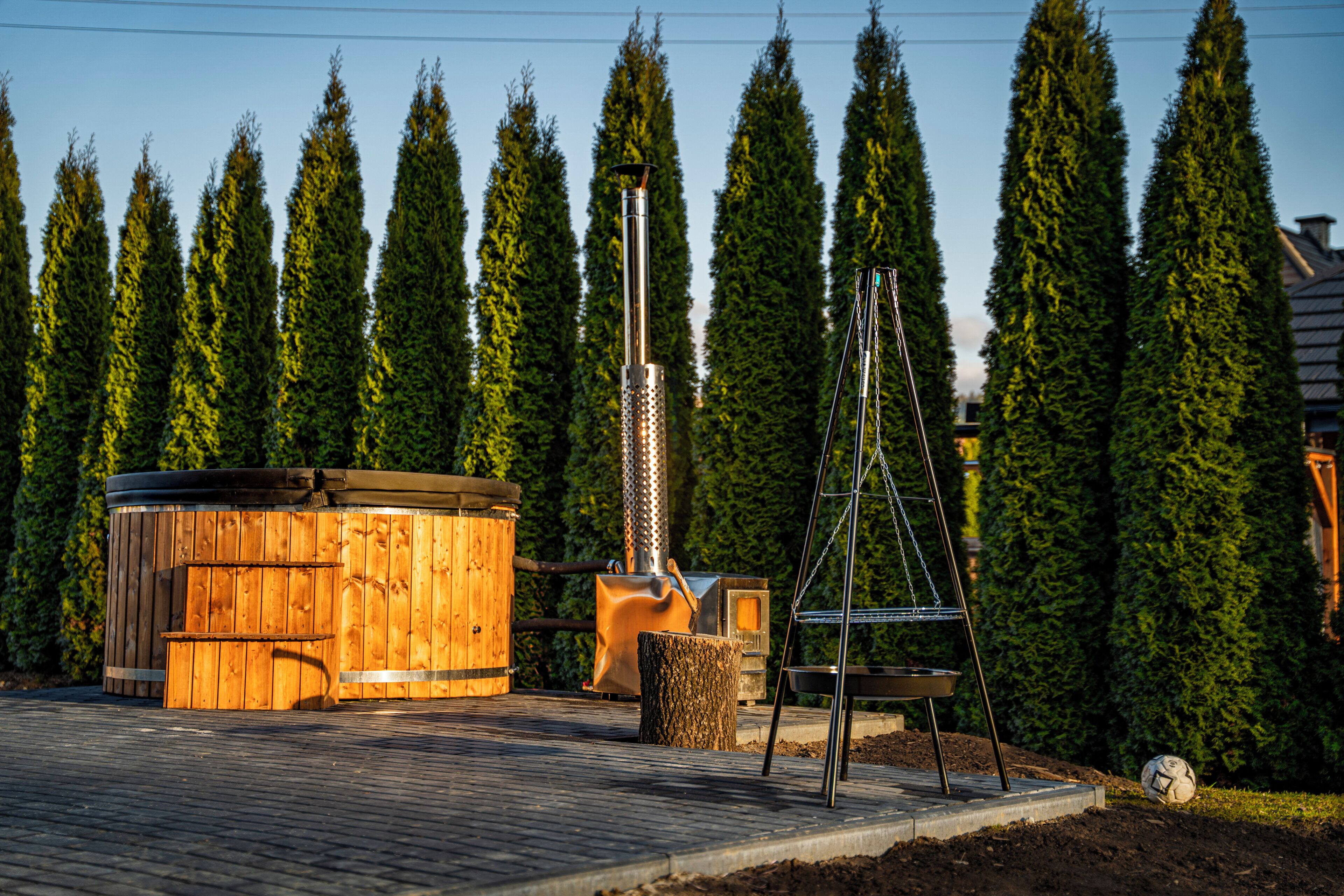 A wooden hot tub, a metal structure, and a soccer ball on a paved area with tall trees in the background.