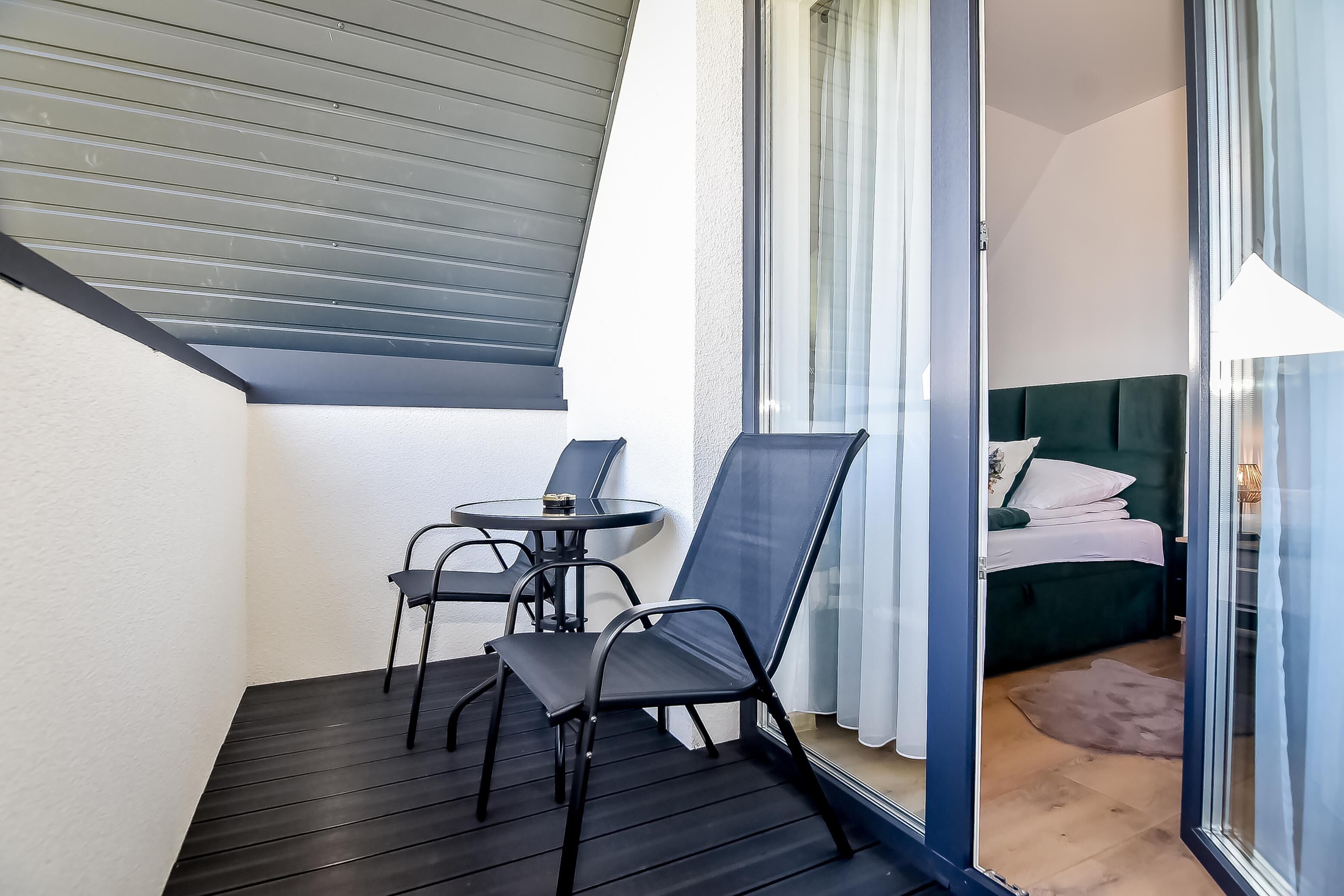 A balcony with a table and chairs, a bed with a green headboard, and a view through a glass door.