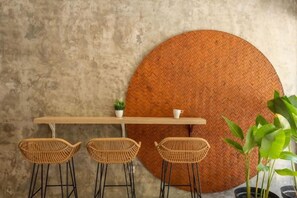A round woven table with four wicker chairs, a potted plant, and a wall-mounted shelf with a small plant and a cup. - Cove Kanaya (Bali)