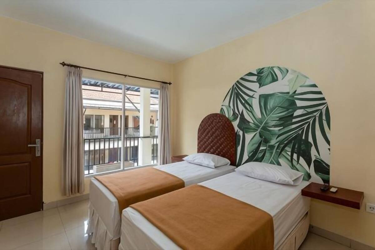 A hotel room with two beds, a large window with curtains, a view of buildings outside, and a wall with a tropical leaf pattern.