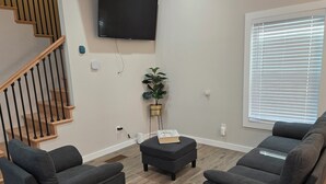 Una sala de estar moderna con un televisor de pantalla plana montado en la pared, una escalera con barandillas de madera y un ventanal con persianas.