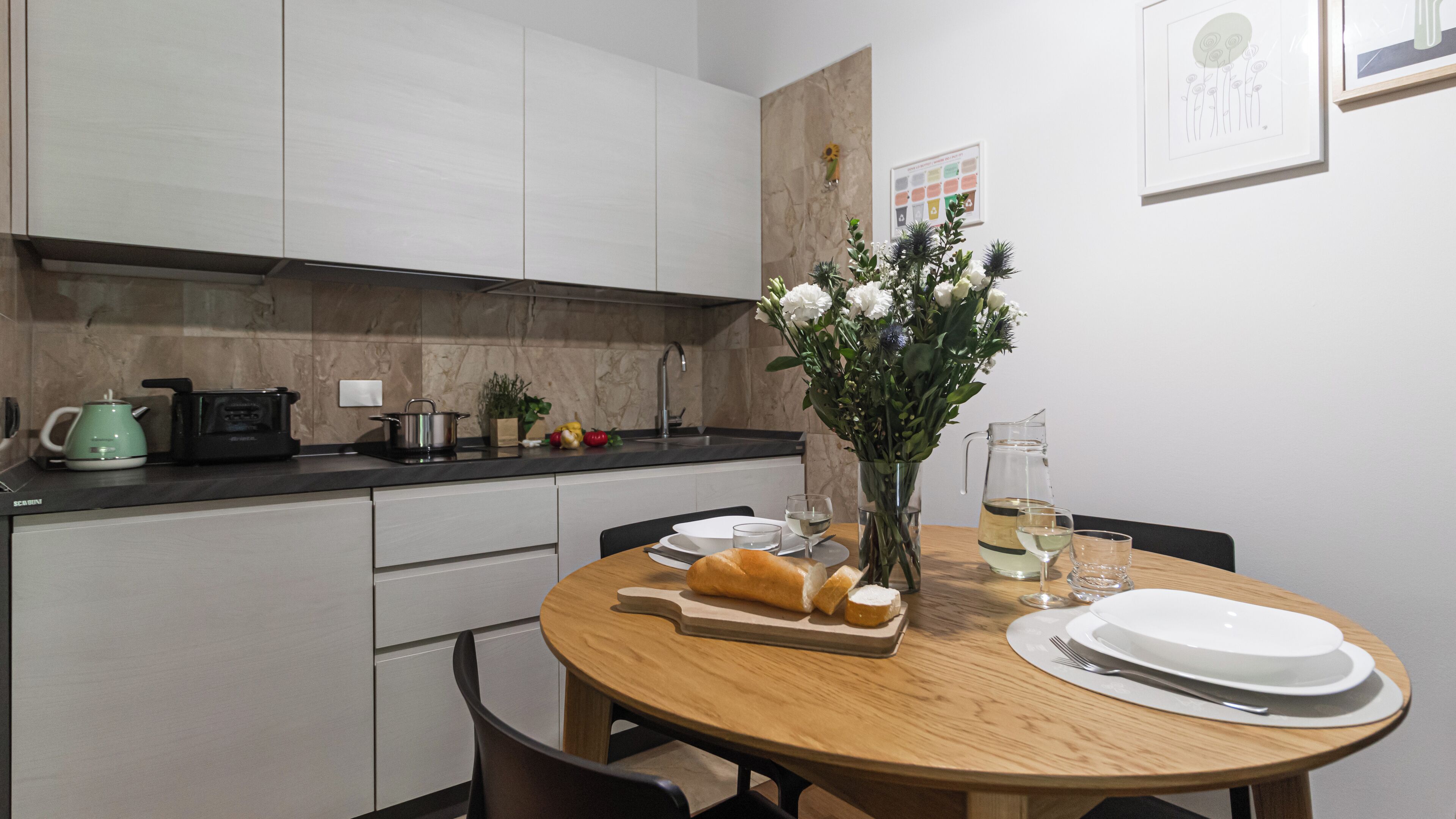 A modern kitchen with white cabinets, a black countertop, and a wooden dining table set with plates, a pitcher of water, and a loaf of bread.