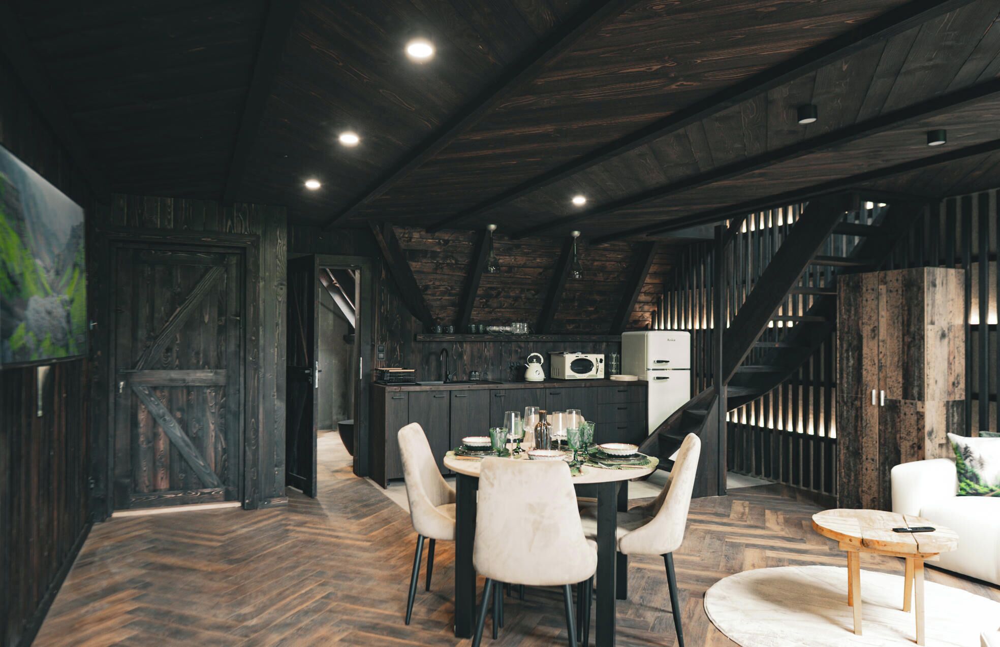 Una cocina rústica con vigas de madera, un comedor con una mesa redonda y sillas, y una escalera que sube a un nivel superior.