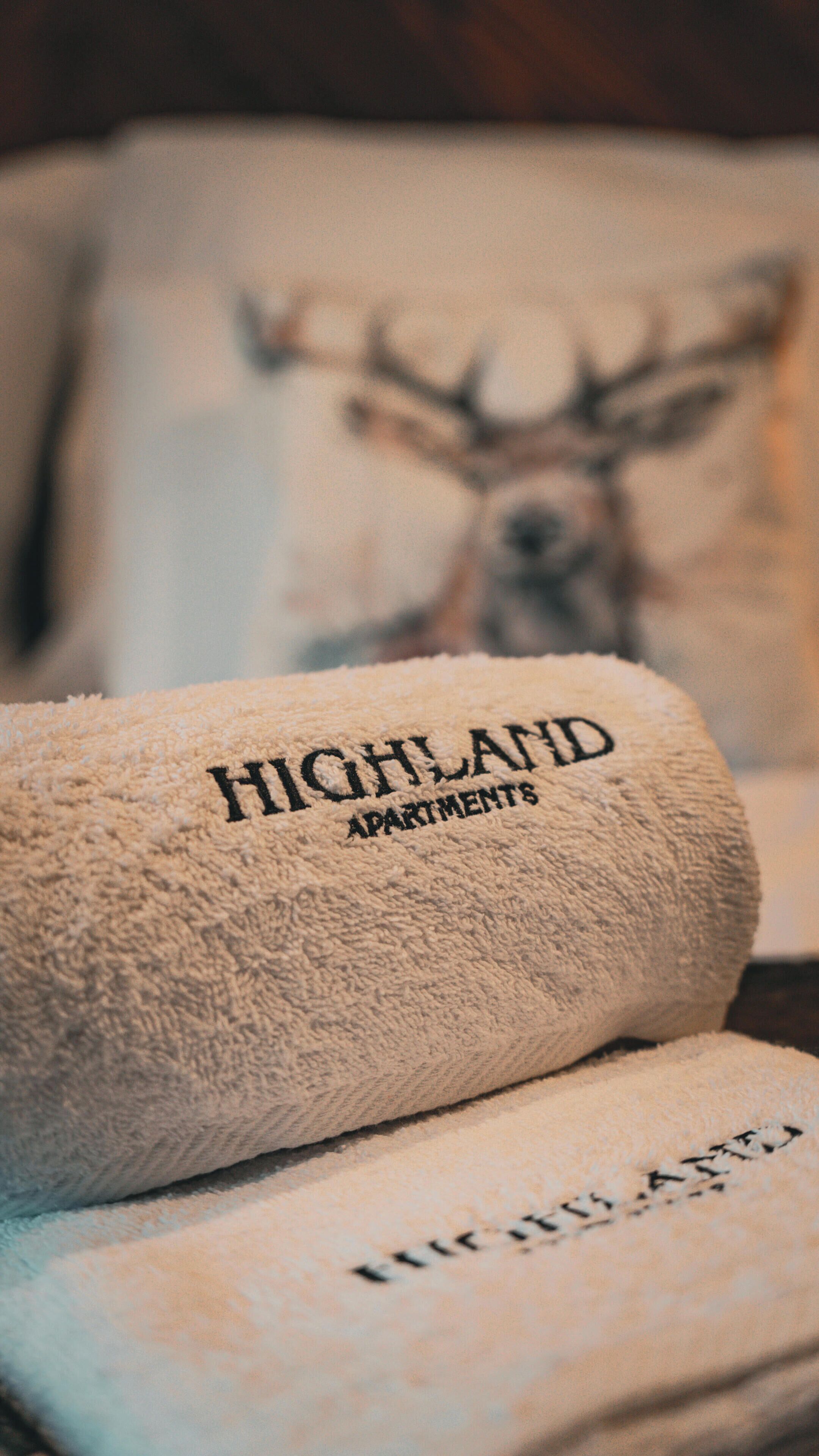 A rolled towel with "Highland Apartments" printed on it, placed on a bed with a pillow featuring a deer image.