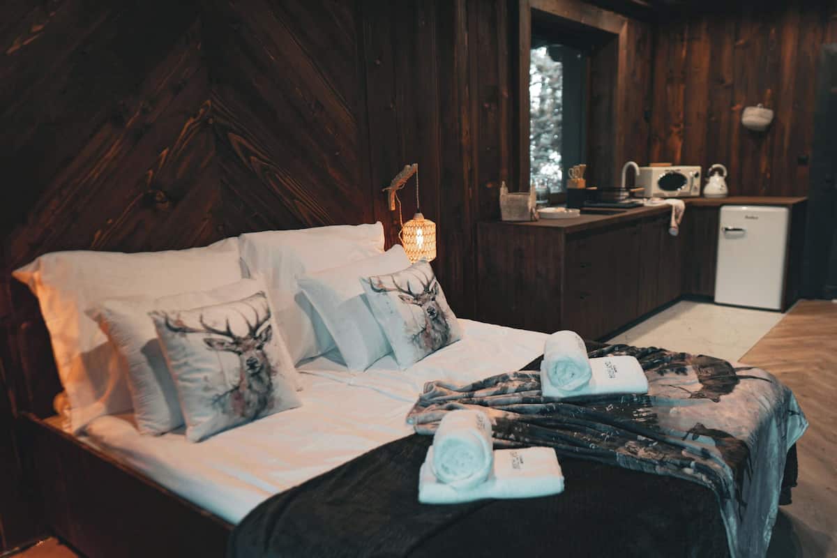 Ein gemütliches Bett mit Hirsch-Motiv-Kissen, ein ordentlich bezogenes Bett mit aufgerollten Handtüchern, eine kleine Küchenzeile mit Spüle und Kühlschrank sowie ein rustikales Holzambiente.
