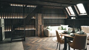 Une pièce chaleureuse de style loft, avec un parquet en bois disposé en motif chevrons, un canapé blanc, une table à manger pour quatre personnes et un escalier doté d’une rampe en bois.