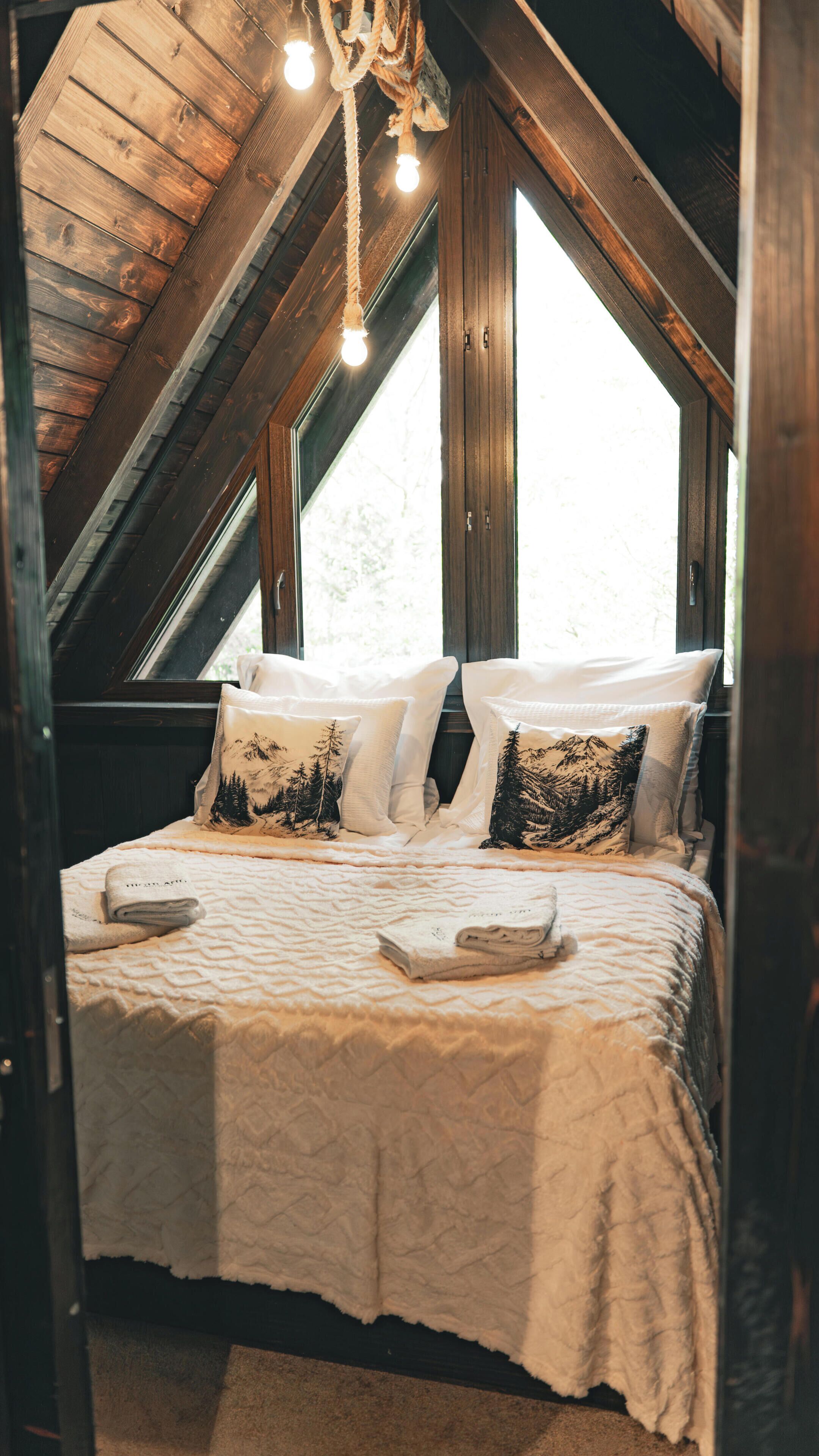 A cozy attic bedroom with a sloped ceiling, wooden beams, and a large window. There is a bed with white bedding and two pillows with forest designs.
