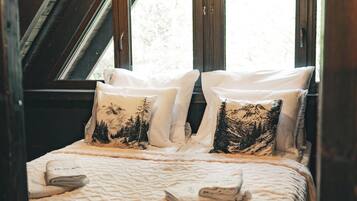A cozy attic bedroom with a sloped ceiling, wooden beams, and a large window. There is a bed with white bedding and two pillows with forest designs.