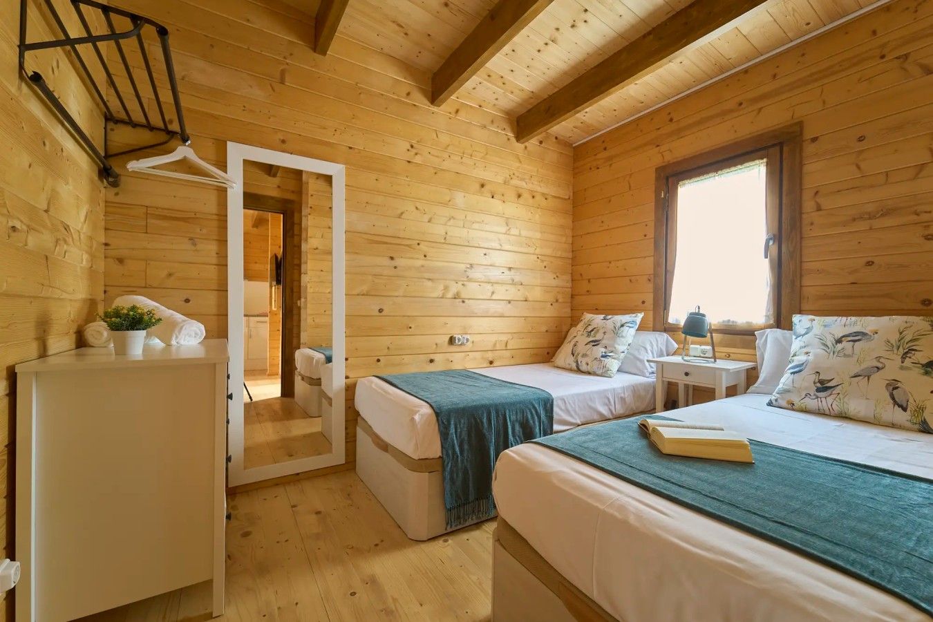A cabin with two beds, a small table, a window, and a door leading to another room.