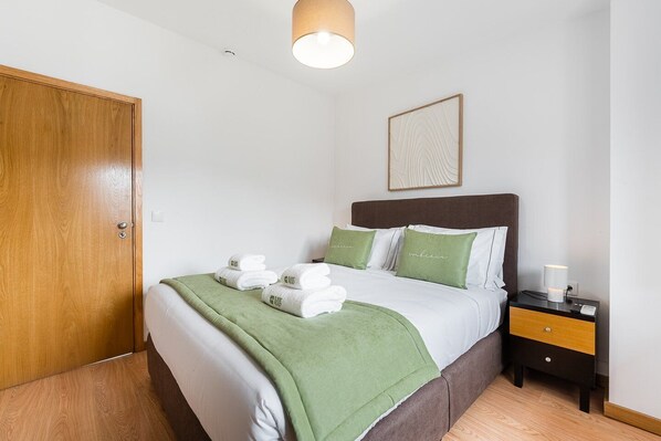 Una cama bien hecha con ropa de cama verde y blanca, una mesita de noche con una lámpara, y una puerta de madera.