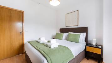 A neatly made bed with green bedding and white towels, a bedside table with a lamp, and a wooden door.