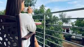 Eine Person sitzt auf einem Stuhl mit Metallgestell, eine Tasse haltend, auf einem Balkon mit Geländer.
