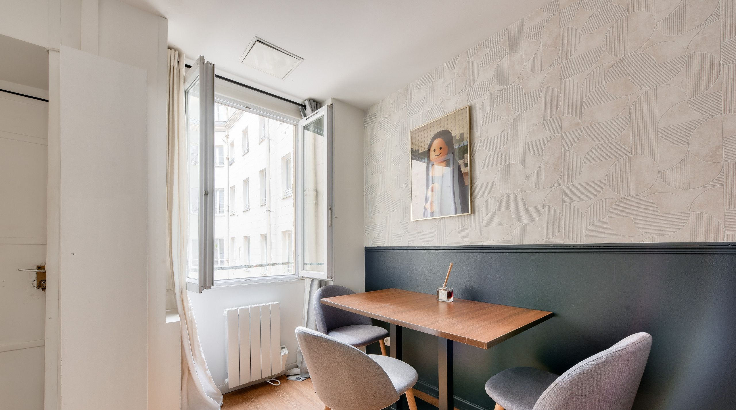 Een moderne eetruimte met een houten tafel en twee grijze stoelen, een raam met gordijnen en een wand met een geometrisch patroon.