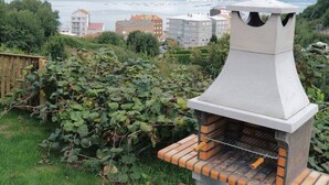An outdoor barbecue grill with a chimney, set in a grassy area with a view of a body of water and buildings in the background. - Cima 1 Cabin in Bueu (Bueu)