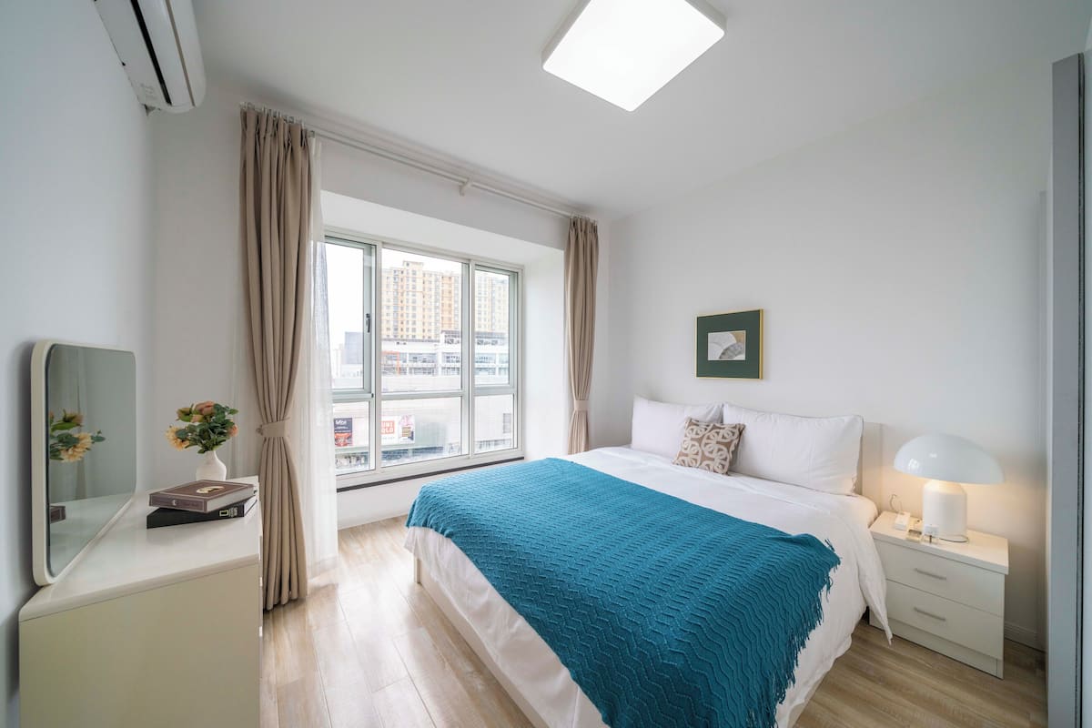 A modern bedroom with a large window, a bed with a teal blanket, a bedside lamp, and a small table with a vase and book.