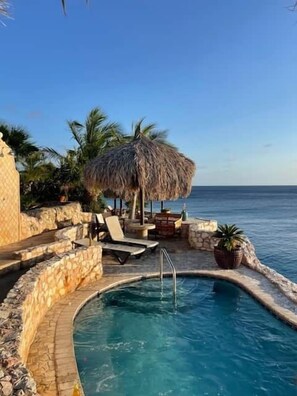 A pool area with lounge chairs and a thatched umbrella, surrounded by stone walls and palm trees.