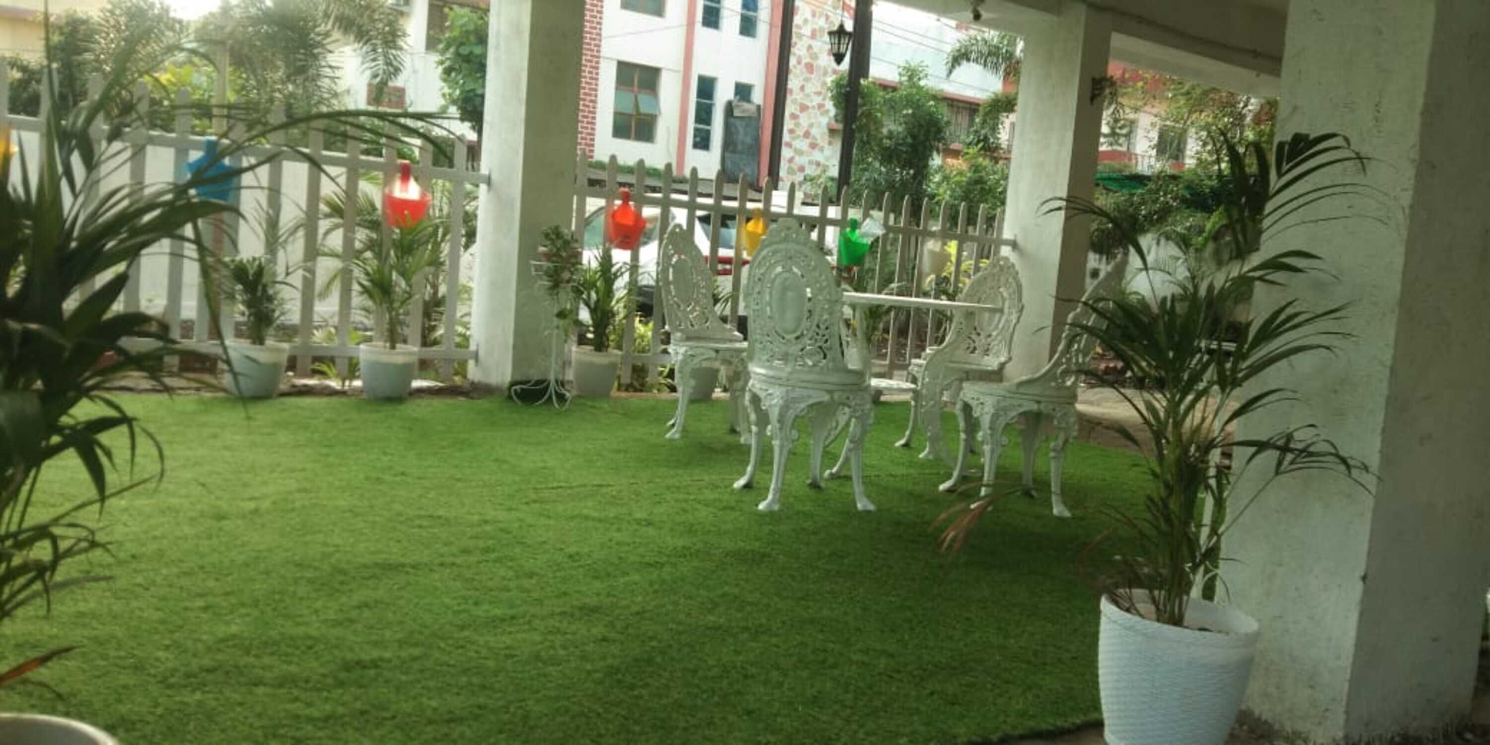 A covered outdoor area with a neatly trimmed green lawn, white metal chairs, and potted plants.