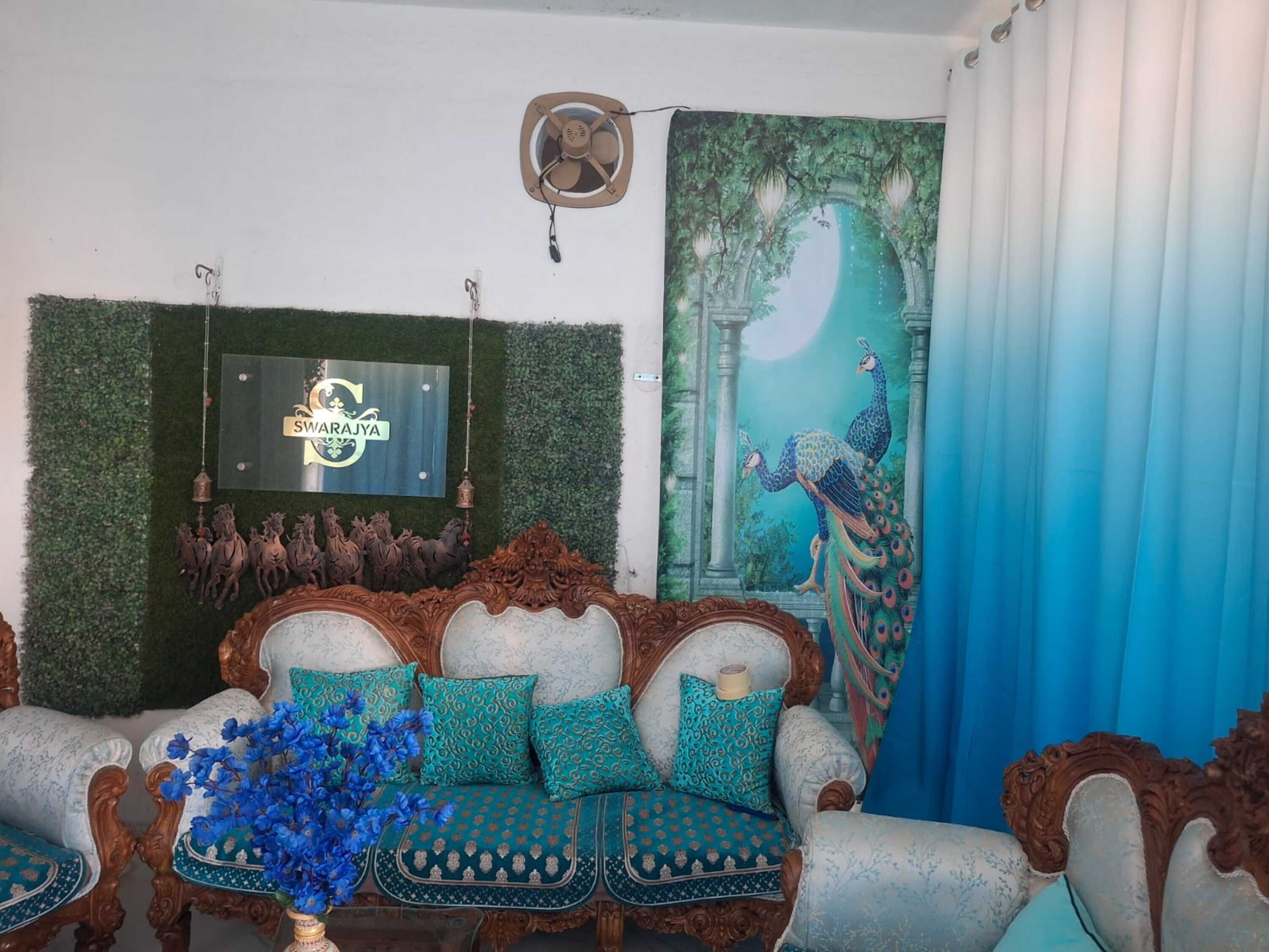 A traditional sofa with ornate woodwork and patterned cushions, a decorative wall with a peacock painting, and a wall-mounted fan.