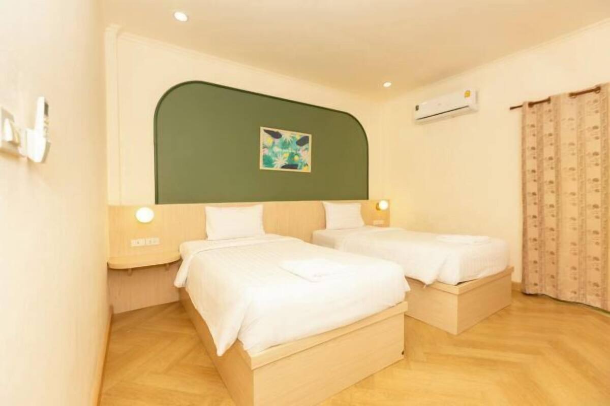 A hotel room with two single beds, a green headboard, a small painting, an air conditioning unit, and a patterned curtain.